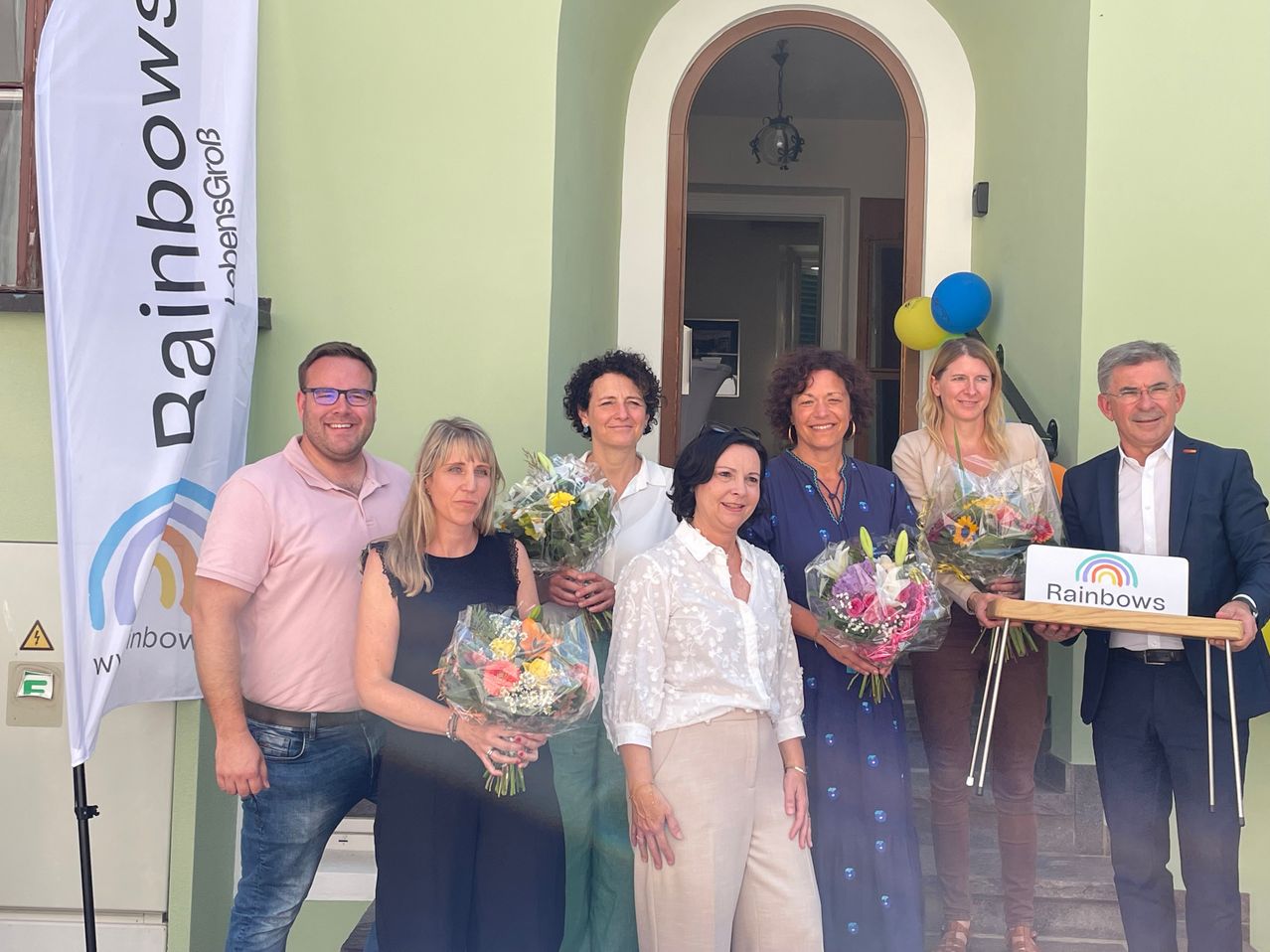 Eine Gruppe lächelnder Erwachsener in verschiedenen Outfits posiert mit Blumen vor einem Gebäude mit einem Banner und Luftballons.