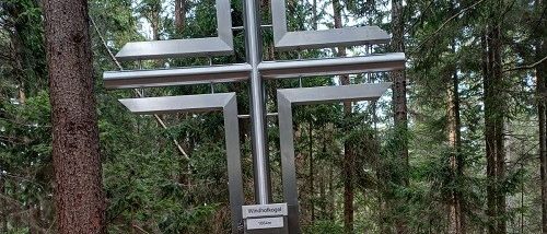 Ein metallisches Kreuzmonument steht auf einem Felsen in einem bewaldeten Gebiet. Es hat eine Tafel mit fremdem Text.