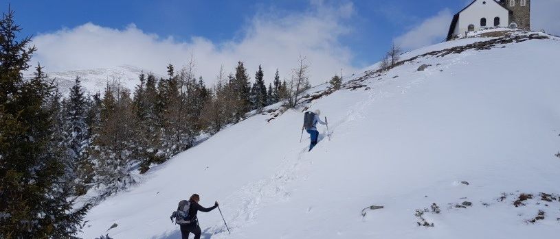 Zwei Wanderer erklimmen einen schneebedeckten Hang mit einer Kirche im Hintergrund. Einer trägt einen Rucksack und Skistöcke.
