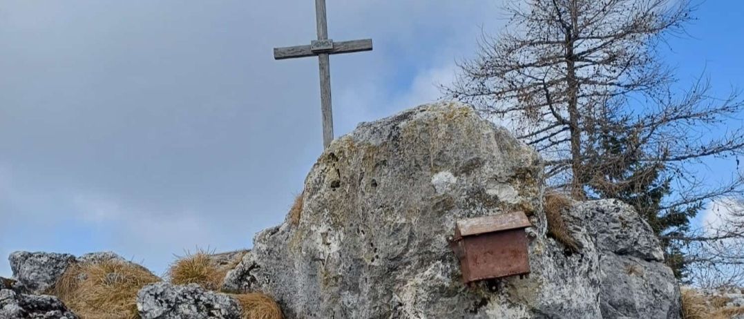 Ein Holzkreuz steht auf einem großen Felsen, mit einer kleinen Metallbox daran befestigt. Der Hintergrund zeigt felsige Hügel und einen klaren blauen Himmel.