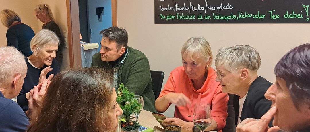 Mehrere Personen sitzen an einem Tisch. Das Speisenangebot auf der Tafel umfasst eine Vielzahl von Frühstücksoptionen, darunter Brot, Tomaten und Marmelade. Sie trinken Wein und scheinen sich in einer entspannten und fröhlichen Stimmung zu befinden.