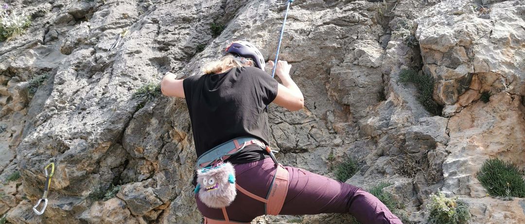 Eine Frau in einem schwarzen T-Shirt und lila Hosen klettert am Fels. Sie hält ein Seil und hat einen pelzigen Gegenstand am Gürtel.