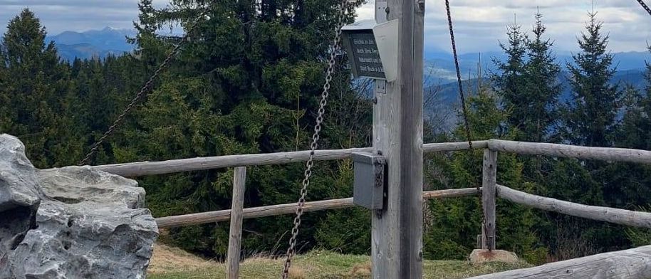 A wooden cross with chains and a metal plaque stands on a mountaintop, surrounded by trees and a wooden fence.