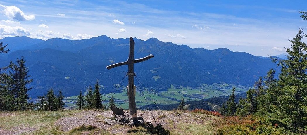 Ein Holzkreuz auf einem Berggipfel mit Blick auf das Tal unten und Berge in der Ferne.