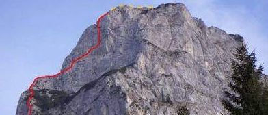 Ein Berggipfel mit einer roten Linie, die eine Kletterroute markiert, umgeben von grünen Bäumen unter einem blauen Himmel.