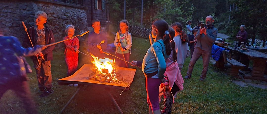 Eine Gruppe von Kindern und Erwachsenen versammelt sich um einen Feuerkorb in einem grün bewachsenen Bereich und röstet Marshmallows. Im Hintergrund sind Bäume und ein Gebäude zu sehen.