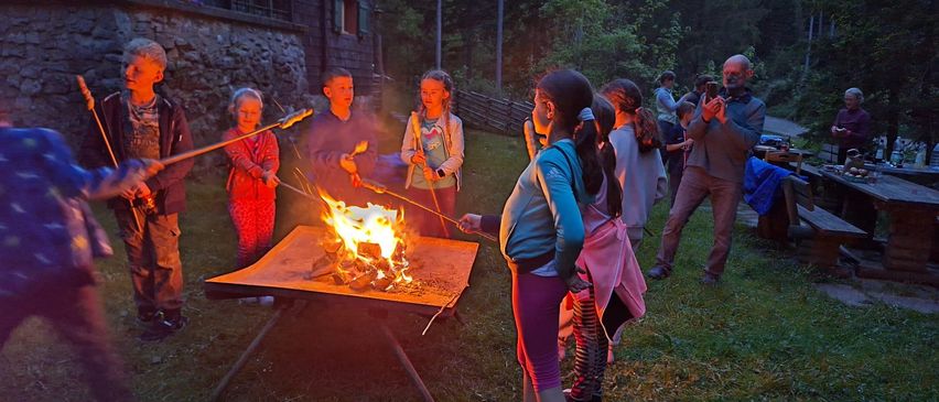 Eine Gruppe von Kindern und Erwachsenen versammelt sich um einen Feuerkorb in einem grün bewachsenen Bereich und röstet Marshmallows. Im Hintergrund sind Bäume und ein Gebäude zu sehen.