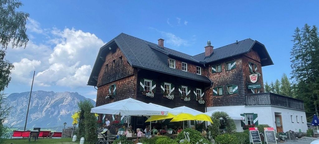 Ein großes Chalet-Gebäude mit Balkon und Schornsteinen. Mehrere Personen sitzen unter Schirmen vor dem Gebäude. Es gibt Pflanzen, einen Kinderwagen und ein geparktes Auto davor. Berge sind in der Ferne.