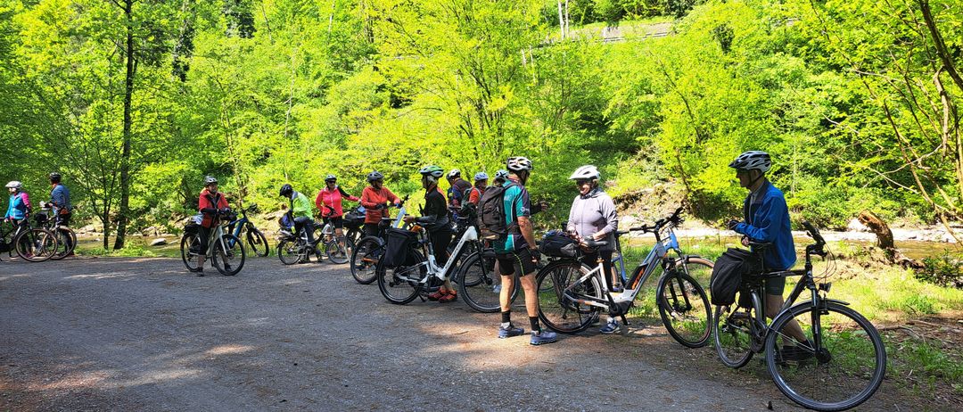 Eine Gruppe von Radfahrern versammelt sich auf einem Kiesweg, umgeben von üppiger Grünfläche. Jeder Radfahrer trägt einen Helm, und einige haben Rucksäcke. Das Gebiet ist gefüllt mit Bäumen und Pflanzen.