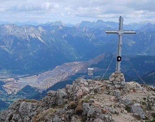Ein Metallkreuz steht auf einem felsigen Berggipfel und blickt auf ein Tal mit Bergen in der Ferne.