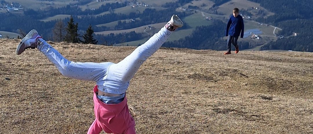Eine Person macht einen Handstand auf einem grasigen Hügel mit Bergen im Hintergrund. Eine weitere Person geht in der Nähe vorbei.