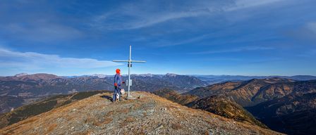 Eine Person in einer blauen Jacke und orangem Hut steht auf einem Berggipfel mit einem Kreuz und einem klaren blauen Himmel im Hintergrund.