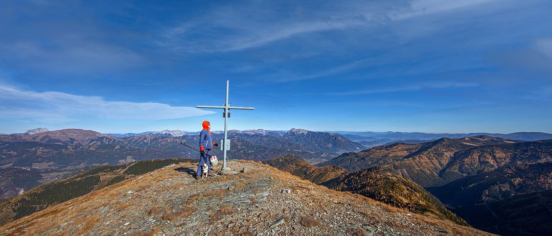 Eine Person in einer blauen Jacke und oranger Mütze steht auf einem Berggipfel mit einem Kreuz und einem Hund, unter einem blauen Himmel mit Wolken.