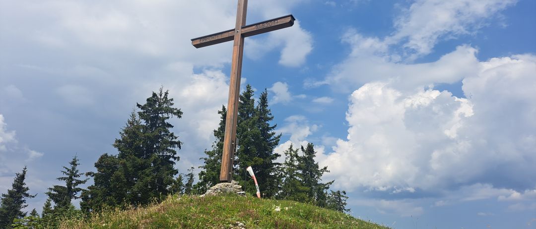 Ein hölzernes Kreuz steht auf einem grasbewachsenen Hügel mit Bäumen im Hintergrund. Das Kreuz trägt das Datum 10.6.2011 und den Namen 'ERRICHTET'. An der Basis des Kreuzes ist eine Flagge platziert.
