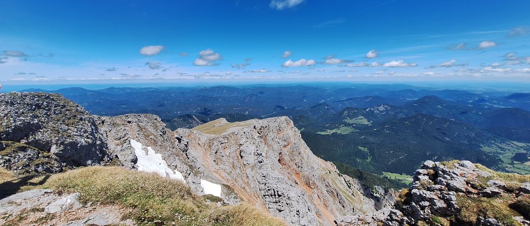 Ein Panoramablick von einem Berggipfel zeigt eine weite Landschaft mit sanften Hügeln und einem blauen Himmel, der von Wolken durchsetzt ist.