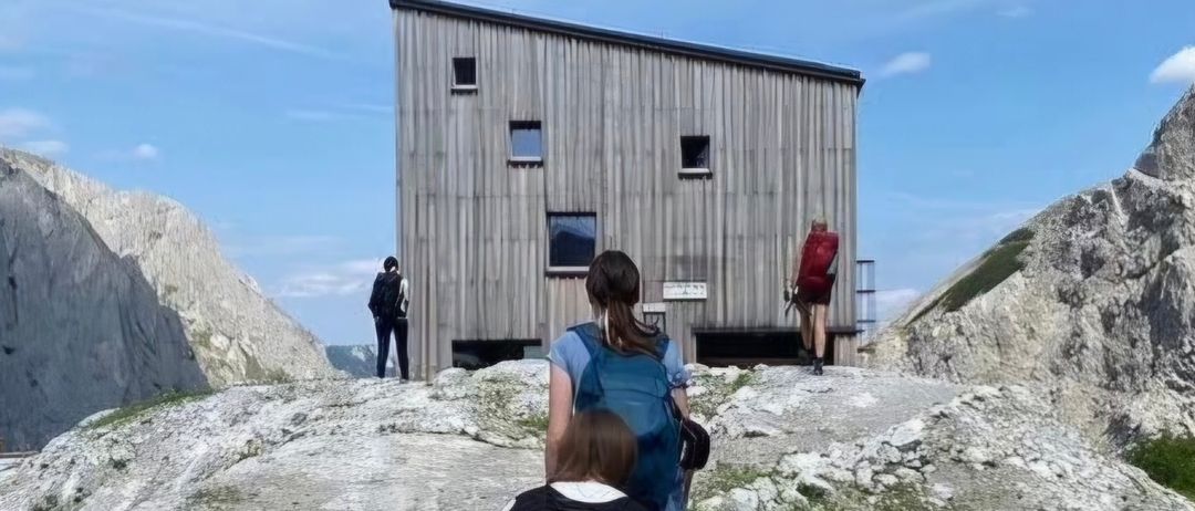 Drei Wanderer mit Rucksäcken erklimmen einen felsigen Pfad in den Bergen, mit einer Holzhütte im Hintergrund.