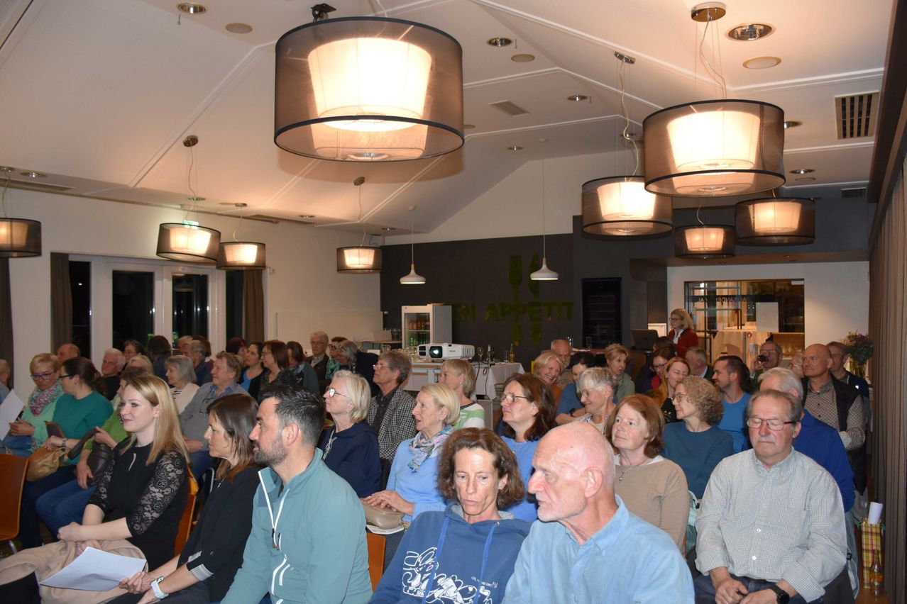 Ein Raum voller Menschen, die sitzen und stehen, einige mit Brille, nehmen an einer Veranstaltung teil. Der Raum ist durch hängende Lampen beleuchtet.
