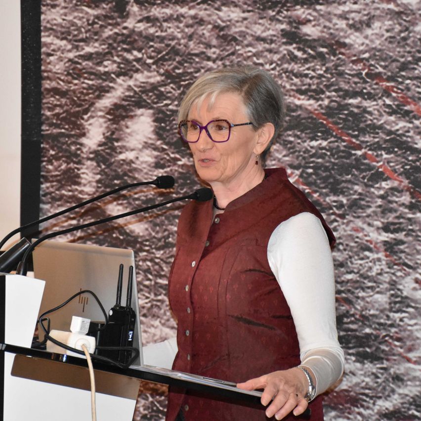 Eine ältere Frau steht an einem Rednerpult und spricht in ein Mikrofon. Sie trägt eine Brille und eine burgunderfarbene Weste über einem weißen Hemd. Ein Laptop und verschiedene Kabel liegen auf dem Pult vor ihr.