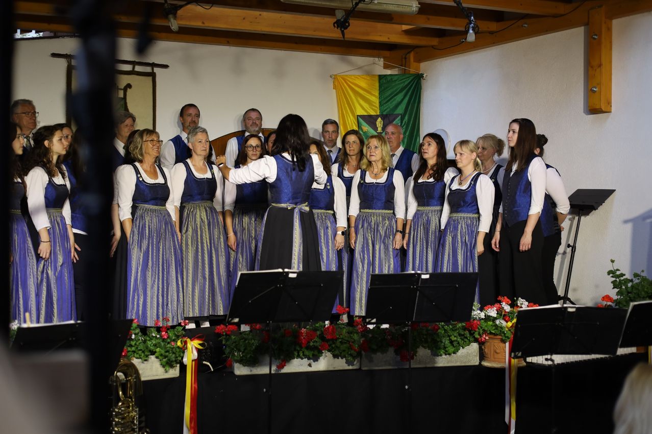 Ein Chor tritt auf der Bühne auf, vor einer Flagge, eine Frau gestikuliert, während andere zusehen, mit Musikinstrumenten und Blumen darunter.
