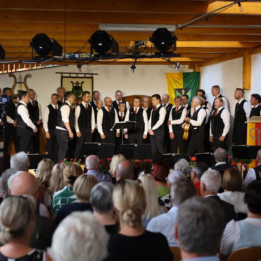 Ein Männerchor in Westen und Krawatten singt auf der Bühne vor einem Publikum, mit Fahnen und Bannern im Hintergrund.