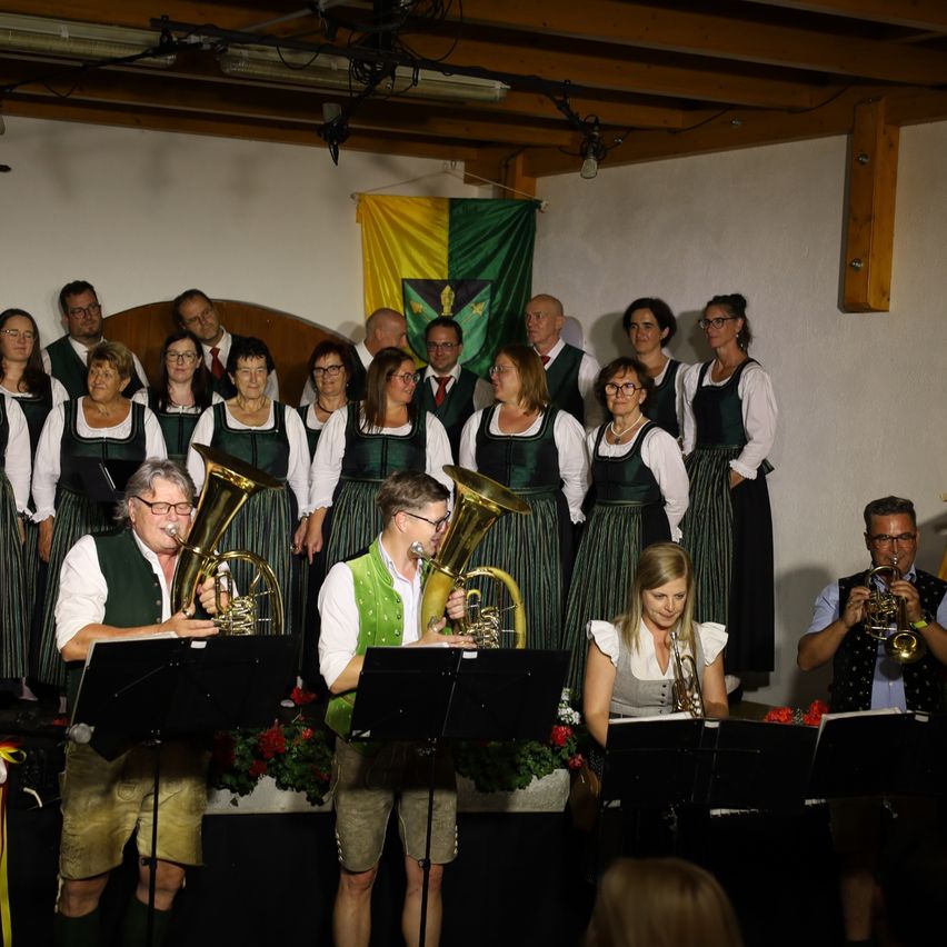 Eine Gruppe von Musikern in traditioneller Kleidung steht auf der Bühne und spielt Blechblasinstrumente. Ein Banner hängt hinter ihnen, und sie werden von Bühnenlichtern beleuchtet.