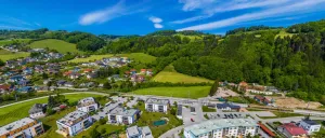 Luftaufnahme eines Bergdorfes mit modernen Gebäuden, grünen Feldern und einem klaren blauen Himmel mit Wolken. Das Dorf liegt eingebettet in den Bergen, umgeben von üppiger Grünfläche.