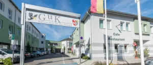 Ein großes weißes Gebäude mit dem Schild 'GUK Rabenstein an der Pielach'. Ein Fahnenmast steht rechts. Mehrere Autos sind auf dem Parkplatz geparkt. Der Himmel ist klar und blau.