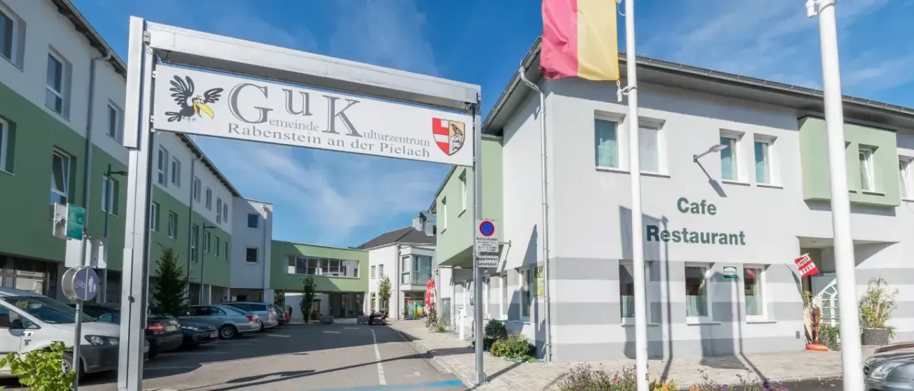 Ein großes weißes Gebäude mit dem Schild 'GUK Rabenstein an der Pielach'. Ein Fahnenmast steht rechts. Mehrere Autos sind auf dem Parkplatz geparkt. Der Himmel ist klar und blau.