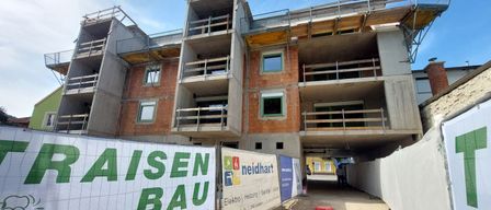 Eine Baustelle mit einem im Bau befindlichen Gebäude, das mehrere Stockwerke, Balkone und ein Schild mit der Aufschrift 'AISEN BAU' aufweist. Die Baustelle ist von Baumaterialien und Absperrungen umgeben.