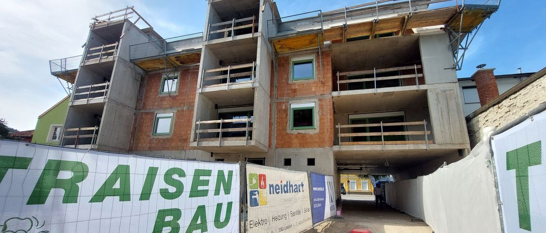 Baustelle mit einem mehrstöckigen Gebäude im Bau, mit Balkonen, Fenstern und einem Schild, das 'AISEN BAU' besagt. Eine Person geht in der Ferne.