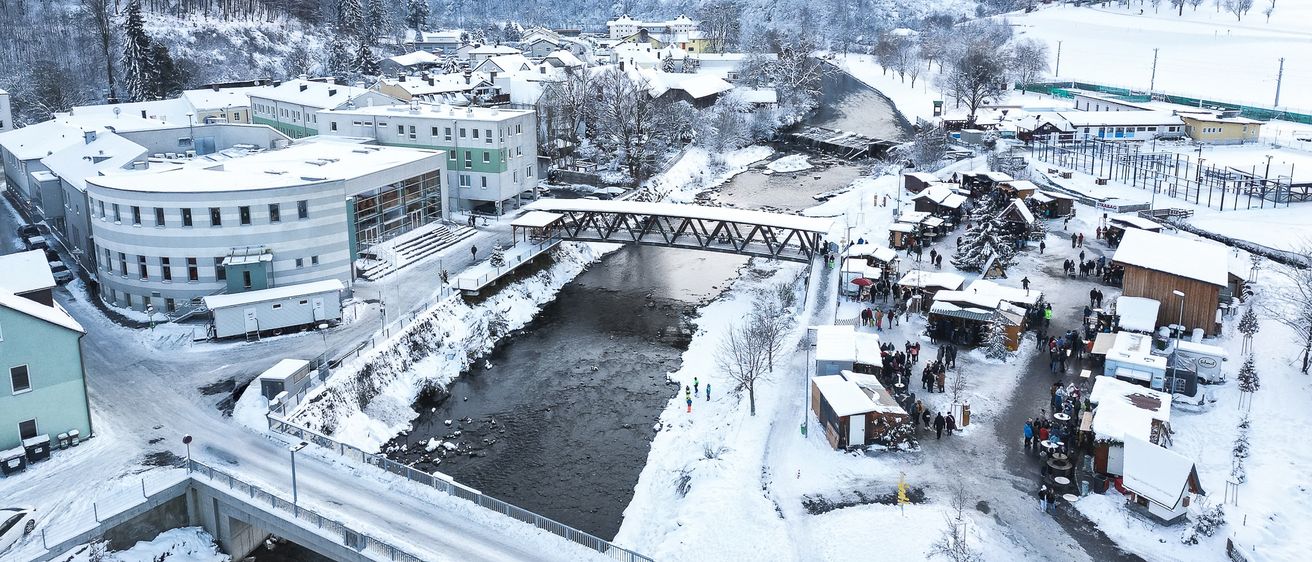 Eine Luftaufnahme einer verschneiten Stadt mit einem Fluss, der durch sie fließt. Gebäude sind schneebedeckt, und Menschen sind in der Nähe einer Brücke und bei Marktständen zu sehen. Die Berge im Hintergrund sind ebenfalls schneebedeckt.
