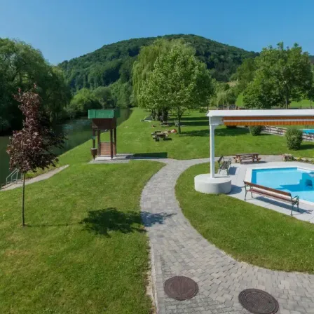 Eine Draufsicht auf einen Park am Flussufer mit einem Pool, einem kleinen Pavillon und Picknicktischen unter Bäumen. Ein Fluss fließt neben dem Gras.