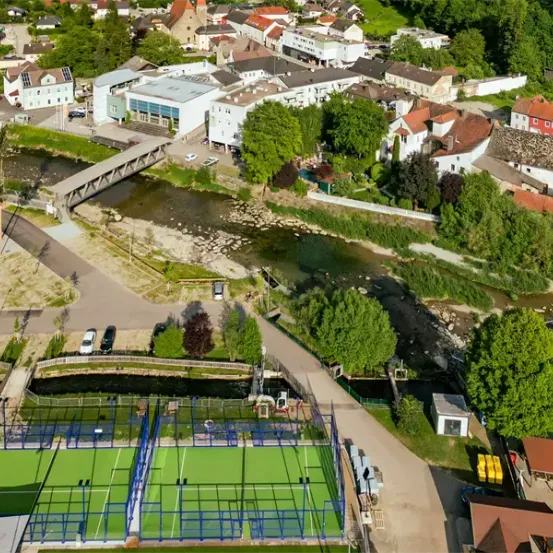 Eine Luftaufnahme einer Stadt mit einem Fluss, Häusern und einem Tennisplatz in der Nähe einer Brücke. Bäume und Gebäude säumen die Flussufer.