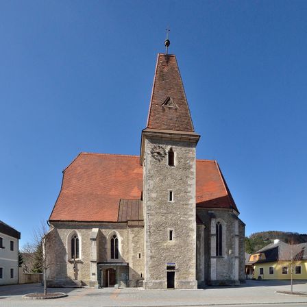 Eine alte Steinkirche mit einem roten Dach und einem Kirchturm mit einer Uhr. Sie befindet sich auf einem Platz in der Stadt mit einem gelben Gebäude auf der rechten Seite.