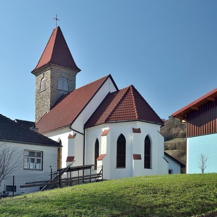Eine weiße Kirche mit einem braunen Dach und einem Turm, unter einem klaren blauen Himmel. Sie ist von einem grasigen Hügel umgeben und hat ein kleines Gebäude rechts neben sich.