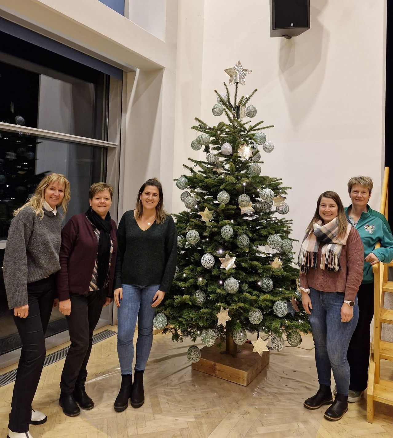 Fünf Frauen stehen neben einem Weihnachtsbaum und lächeln für ein Foto. Der Baum ist mit silbernen Kugeln und Sternen geschmückt. Sie befinden sich in einem Raum mit einem Glasfenster.