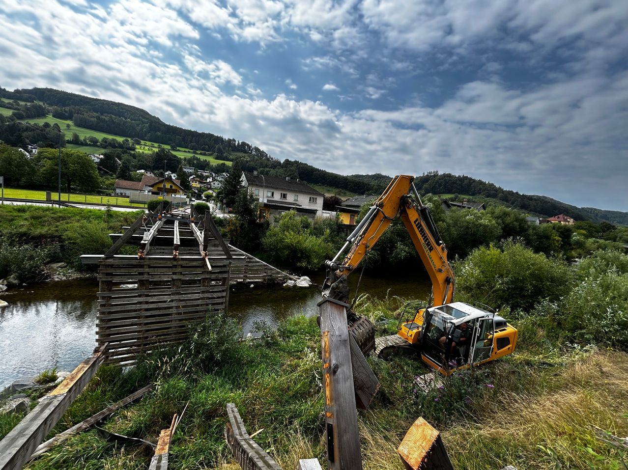 Ein Bauplatz mit einem gelben Bagger, der eine hölzerne Brücke neben einem Fluss entfernt, vor einer Kulisse aus üppiger Grünfläche und Bergen.