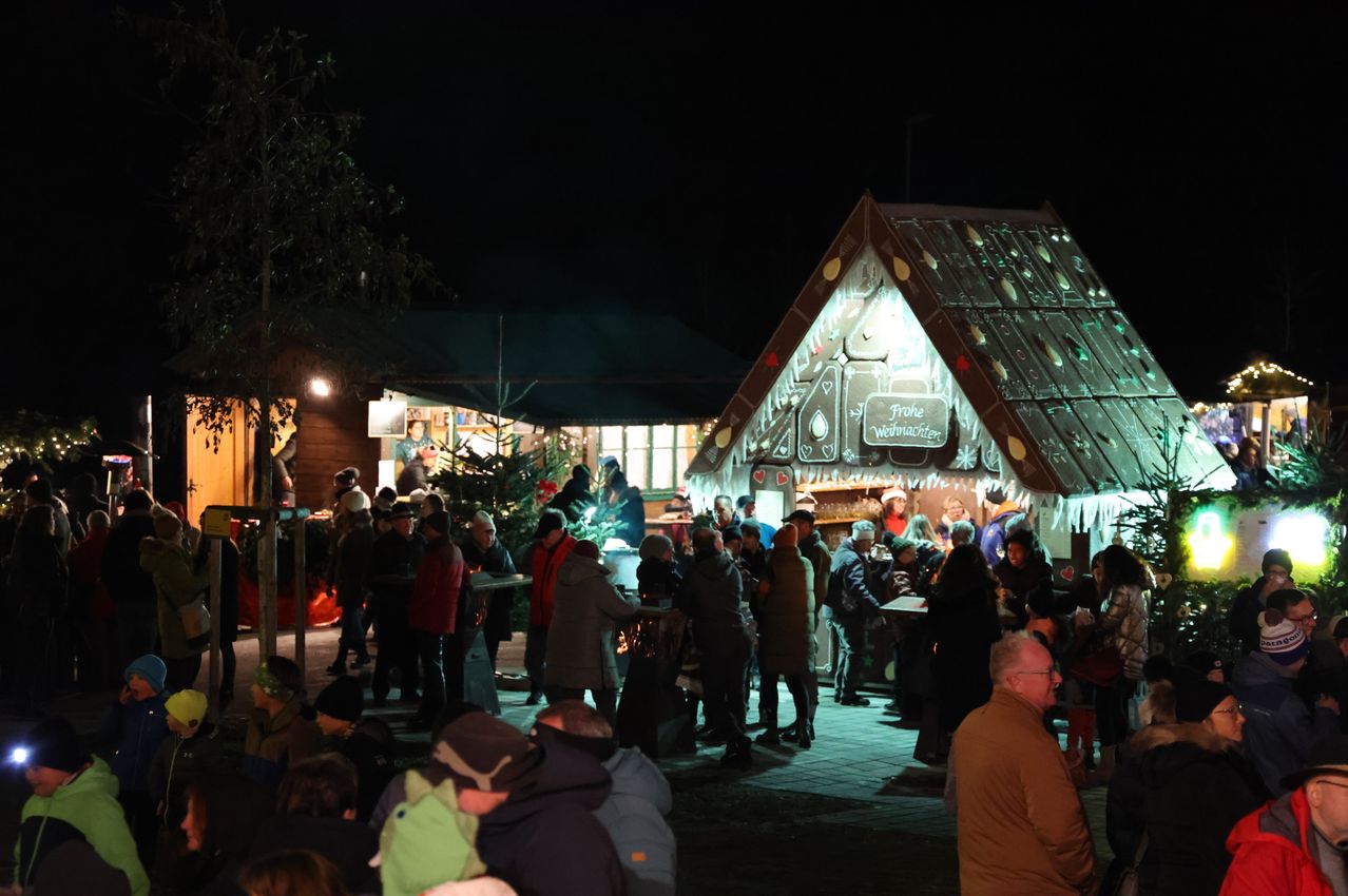 Eine Menschenmenge versammelt sich nachts um ein Lebkuchenhaus. Das Haus ist mit Lichtern dekoriert und das Wort 'Weihnachten' ist darauf geschrieben. Einige Leute stehen und reden, während andere gehen. Eine Straßenlaterne ist links und ein Baum ist dahinter.