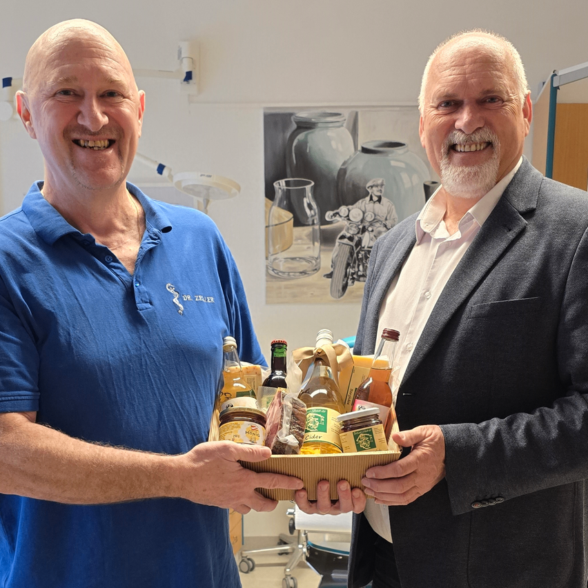 Zwei lächelnde Männer, einer in einem blauen Shirt und der andere in einem grauen Anzug, halten eine Box mit Essen und Getränken.