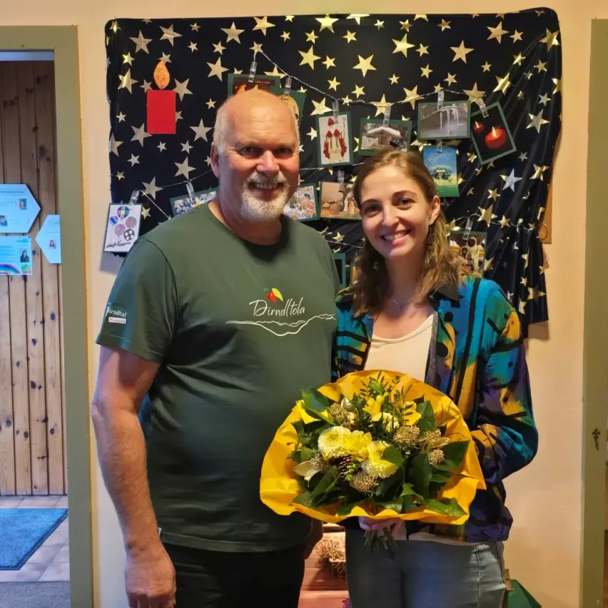 Ein Mann und eine Frau stehen nebeneinander vor einem Hintergrund mit Sternen und einer Kerze. Der Mann trägt ein grünes T-Shirt und die Frau hält einen gelben Blumenstrauß.