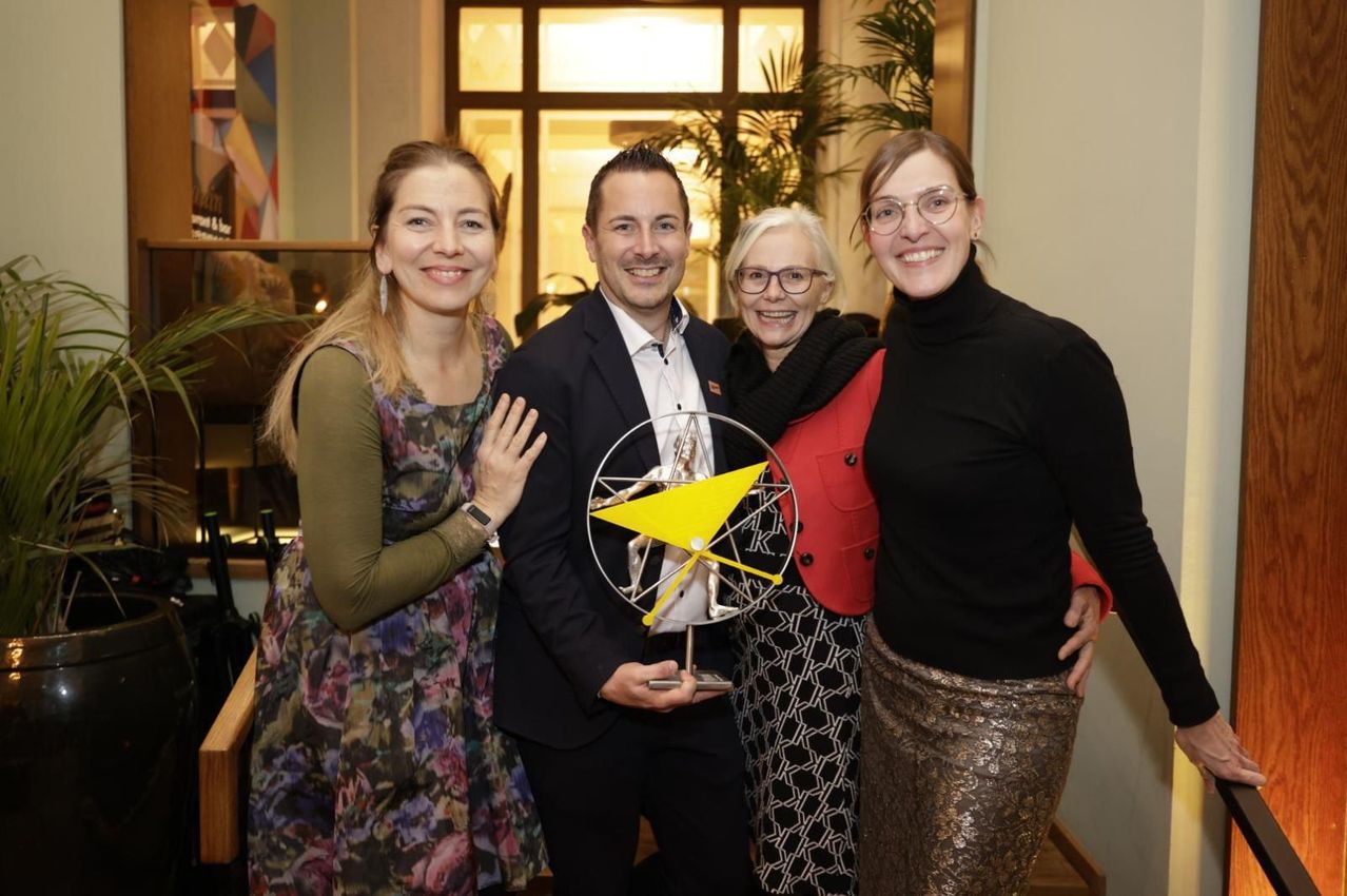 Vier Menschen lächeln und posieren für ein Foto mit einem Mann, der einen Pokal hält. Hinter ihnen ein Glasfenster mit Pflanzen.