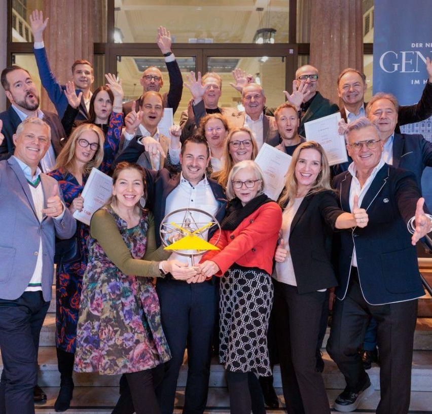 Eine Gruppe von Menschen steht auf einer Treppe, hält Urkunden und eine Trophäe in den Händen und posiert für ein Foto mit Lächeln und Gesten.
