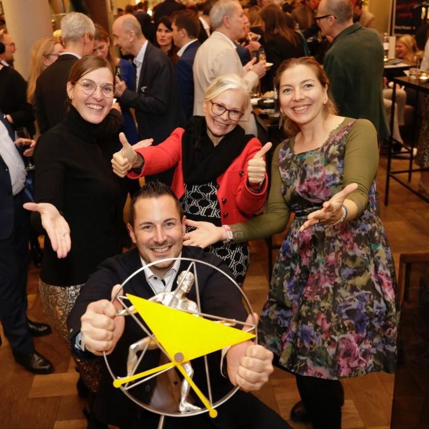 Eine Gruppe lächelnder Menschen bei einer Veranstaltung. Ein Mann in der Mitte hält eine Trophäe, und drei Frauen stehen hinter ihm und geben Daumen hoch.