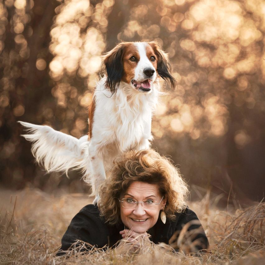 Eine Frau liegt auf dem Gras mit einem Hund auf ihrem Rücken. Sie lächelt und trägt eine Brille. Der Hund schaut nach oben. Der Hintergrund ist verschwommen mit Bäumen.
