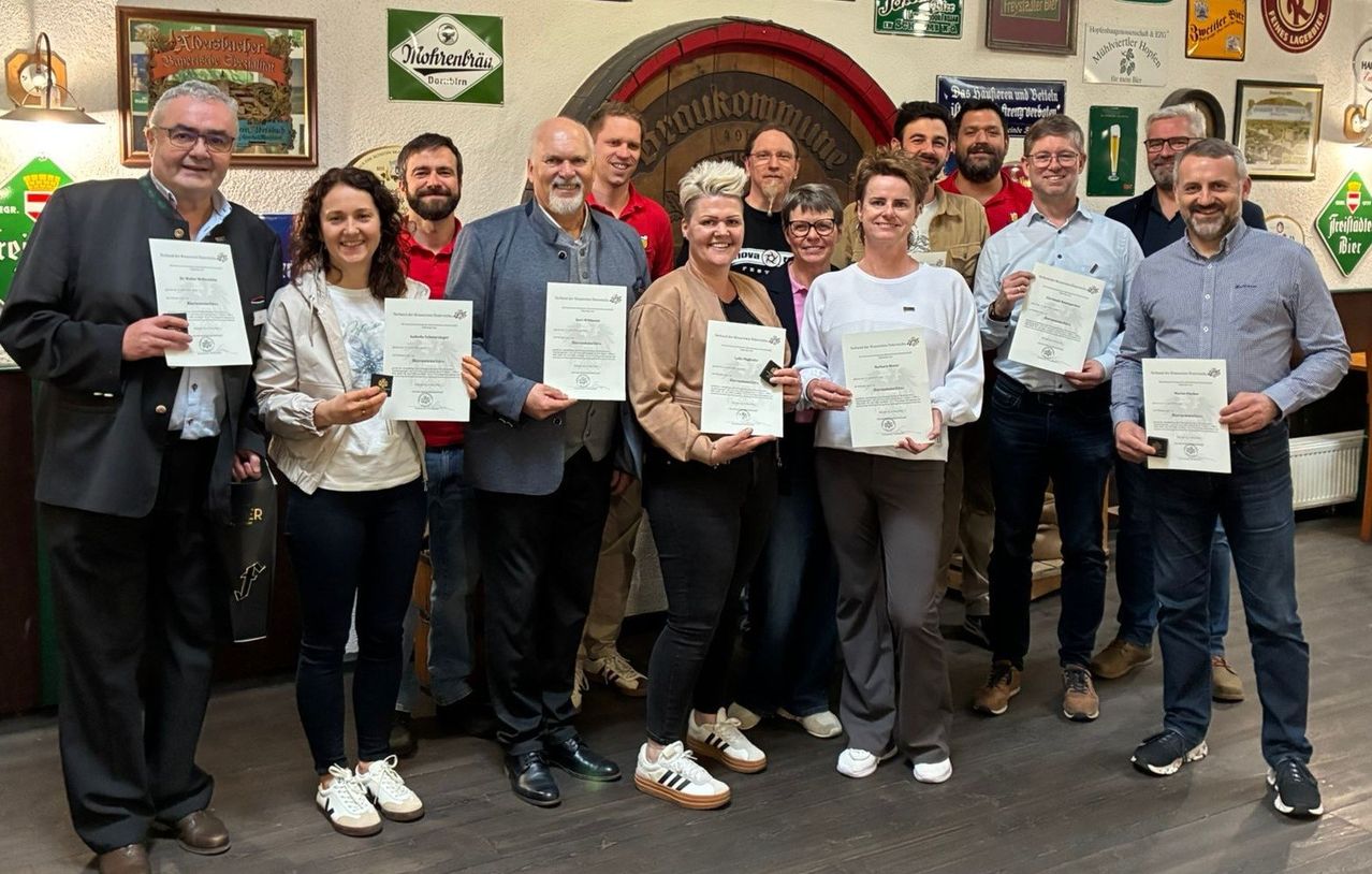 Eine Gruppe von Menschen steht vor einem Holzfass und hält Zertifikate. Sie tragen Freizeitkleidung und lächeln für ein Foto. Hinter ihnen hängen gerahmte Bilder an der Wand.