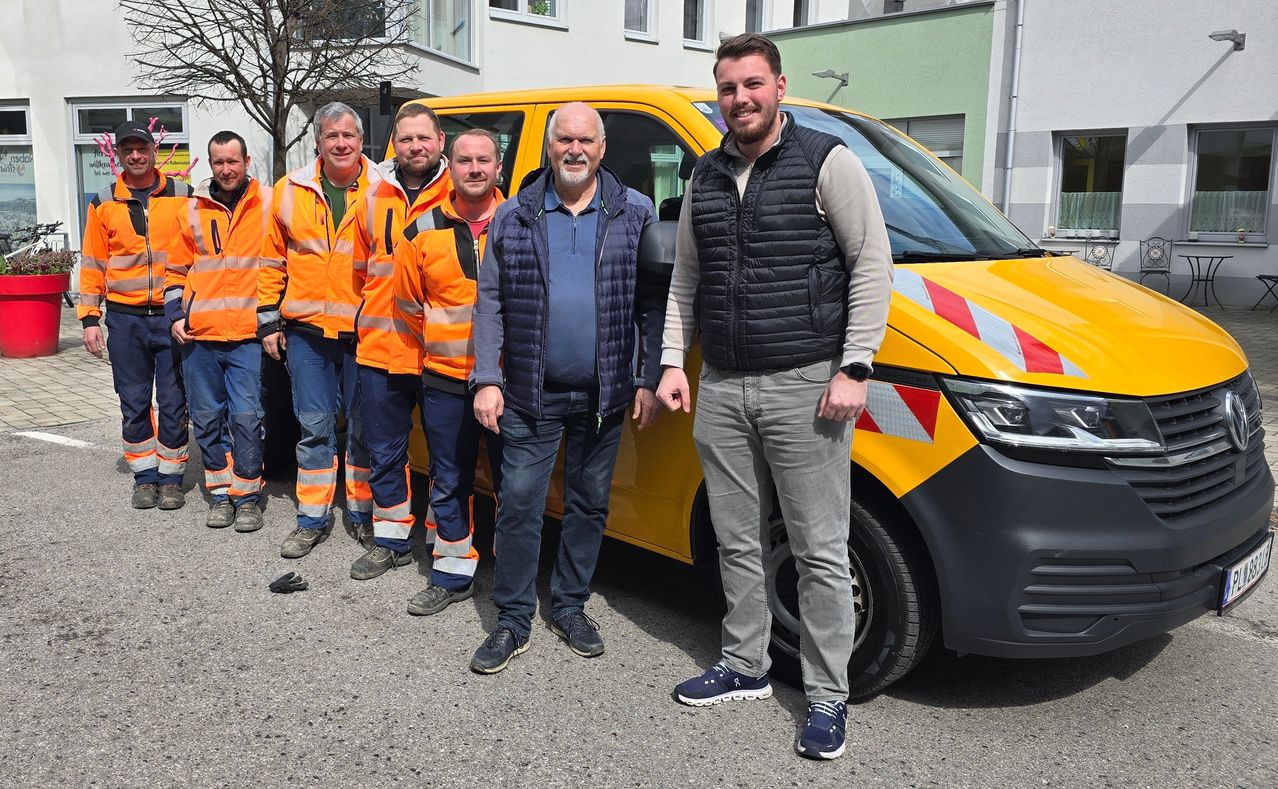 Eine Gruppe von Männern in orangefarbenen und grauen Arbeitswesten steht vor einem gelben Van. Sie scheinen Arbeiter zu sein.