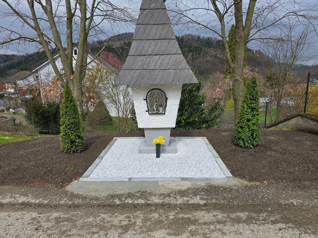 Ein kleines weißes Denkmal mit einem grauen Dach steht in einem Garten. Es hat ein Kreuzdesign auf der Vorderseite und einen gelben Blumentopf darunter. Der Garten ist von Bäumen und einem Zaun umgeben. In der Ferne befinden sich ein Haus und ein Berg.