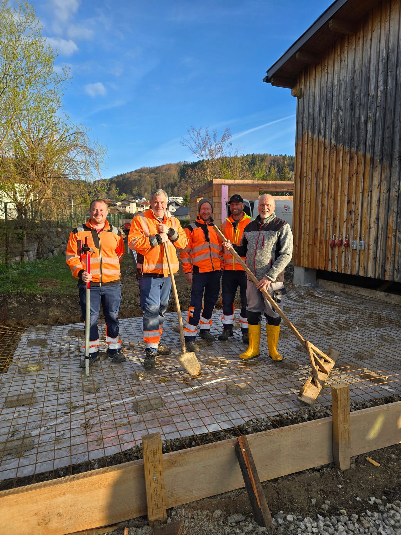 Fünf Männer in Sicherheitswesten posieren mit Werkzeugen auf einem frischen Betonfundament unter einem klaren Himmel.