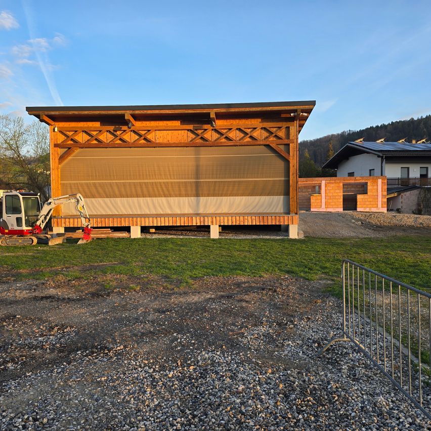 Eine offene Holzbühne mit aufrollbaren braunen Vorhängen, ein Baufahrzeug in der Nähe und ein Haus im Hintergrund unter einem klaren Himmel.