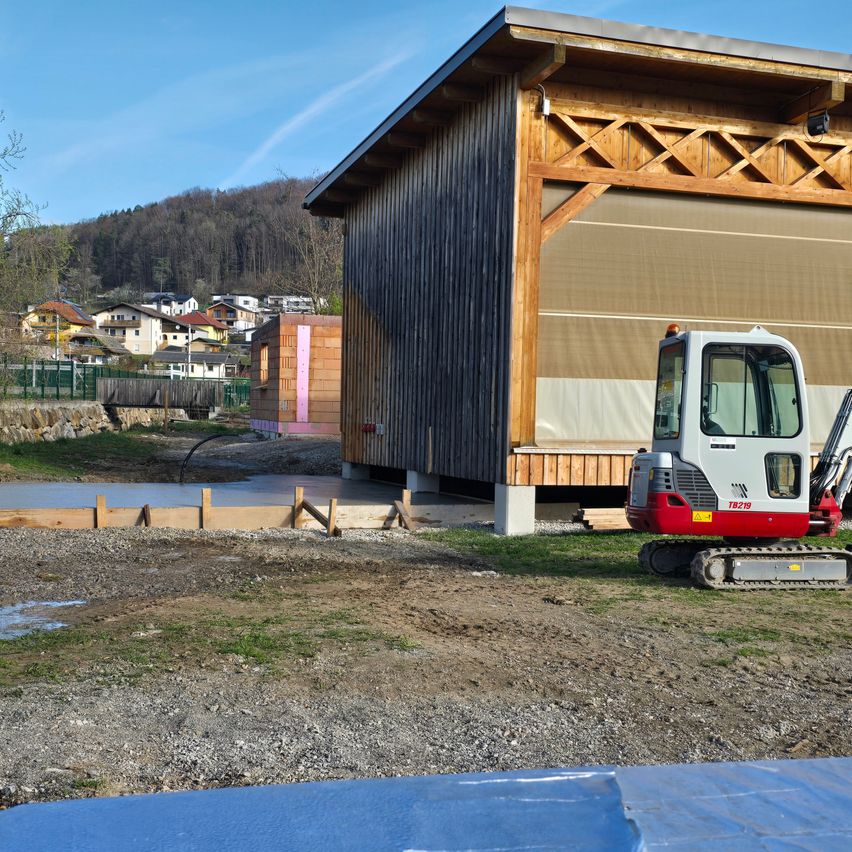 Ein Baustelle mit einem Gebäude im Bau, einem rot-weißen Bagger, einem frisch gegossenen Betonboden und einer Berglandschaft im Hintergrund.